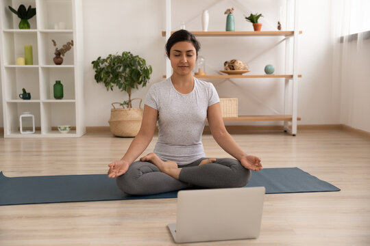 Concentrated Indian Woman Sit On Gymnastic Mat In Lotus Pose Before Laptop Screen Meditate Listen To Calm Music. Peaceful Young Hindu Lady Practice Yoga At Home Learn Asanas From Online Training Class