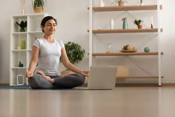 Serene millennial mixed race woman engaged in yoga practice breath deep feel comfort harmony. Tranquil inspired young indian lady sit by laptop on floor at lotus asana meditate to reach peace of mind © fizkes