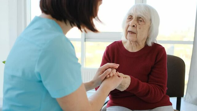 Aged woman talking to nurse while holding hands sitting on chair in nursing home