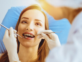 Smiling young woman with orthodontic brackets examined by dentist in sunny dental clinic. Healthy teeth and medicine concept