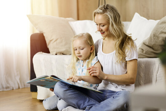 Happy family. Blonde young mother reading a book to her cute daughter while sitting at wooden floor in sunny room. Motherhood concept