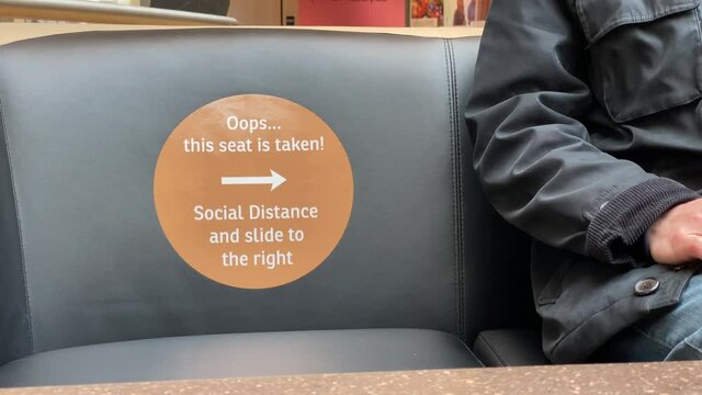 A man sits on a mall couch next to an empty seat with a social distance reminder sticker. Maintaining 6-foot distances while in public spaces was a common sight during the COVID-19 pandemic.  	
