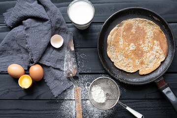 Hot pancake in black pan on black table with flour, milk and eggs