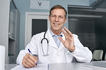 Happy old male doctor waving hand talking to camera during telehealth consultation, consulting patient in virtual meeting, telemedicine e chat. Tele medicine videocall visit, headshot. Webcam view
