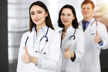 Fototapeta premium Group of young cheerful doctors standing as a team with thumbs up in sunny hospital. Medical help, insurance in health care, best disease treatment and medicine concept