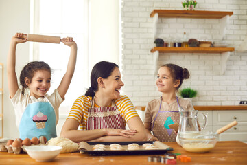 Happy family busy in the kitchen. Young woman and two curious twin daughters in cute aprons cooking together on weekend at home. Mother teaches little children to prepare pastry dough for biscuits