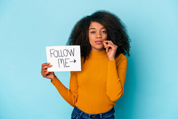 Young african american curly woman holding a follow me placard with fingers on lips keeping a secret.