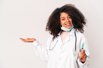 Young african american curly doctor woman holding a syringe showing a copy space on a palm and holding another hand on waist.