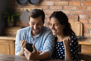 Fototapeta premium Excited millennial Caucasian man and woman feel euphoric win online lottery on smartphone. Happy overjoyed young couple look at cellphone screen triumph with good news or deal message.