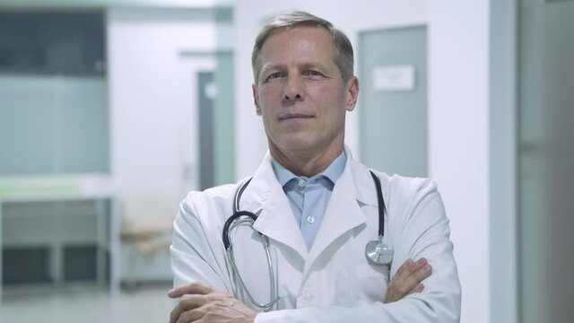 Happy trustworthy proud mature old medical professional doctor stands arm crossed in hospital. Middle aged male senior physician practitioner wears white coat, stethoscope looking at camera. Portrait