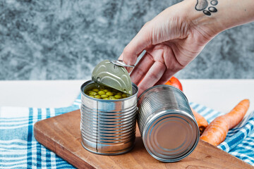 Hand holding a can of boiled green peas on a white table with vegetables and tablecloth