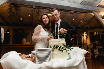 the groom and the bride cut the wedding cake together in a romantic setting. white wedding cake stands on the table. newlyweds are cutting a cake at a banquet. sweet wedding cake decorated 