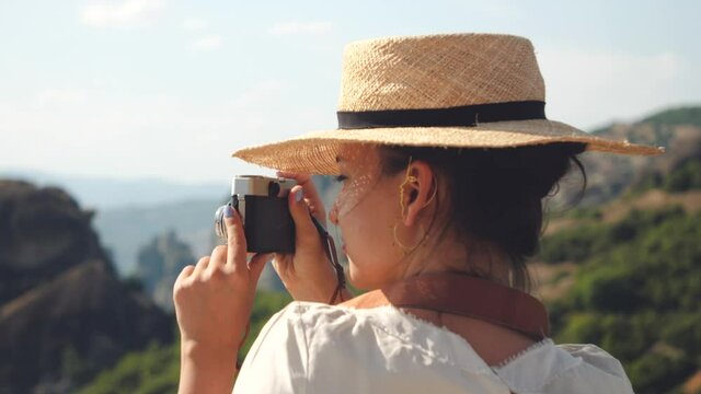 Young woman taking pictures of Meteora rock mountains near Kalambaka, Greece. Vintage camera, large hat, white dress and yellow skirt.