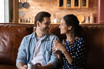 Happy millennial Caucasian couple sit relax on couch at home drink hot coffee talk and chat on domestic weekend. Smiling young man and woman renters rest on sofa enjoy family free time in own house.