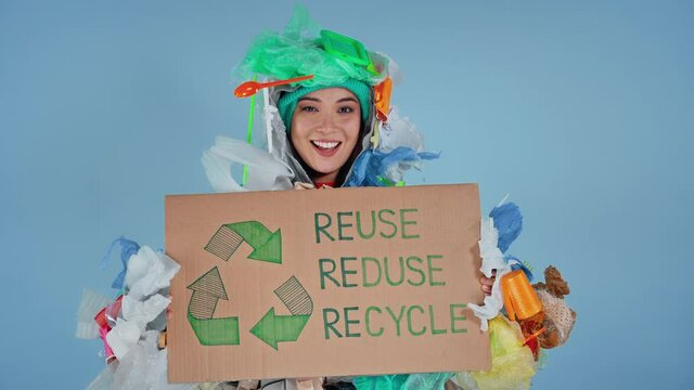 Young asian woman dressed in garbage costume holding cardboard with incription reuse reduse recycle on it. Dressed in the garbage costume and looking at the camera.