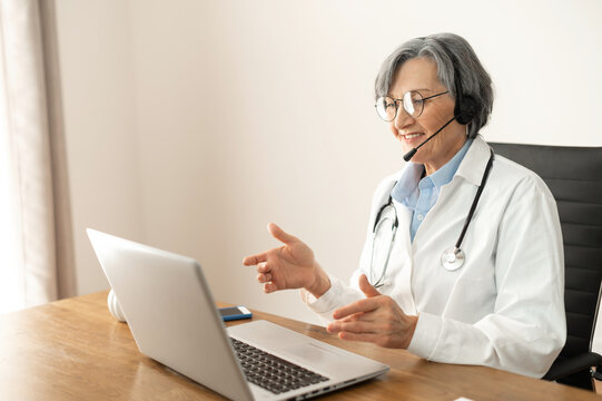 Senior Mature Female Conference Speaker In A Medical Robe, Glasses And A Headset Sitting At The Desk And Teaching Or Talking Online With The Virtual Conference Participants, Healthcare Webinar Concept