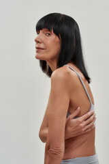 Natural mature beauty. Vertical shot of a senior caucasian woman in lingerie hugging herself, looking at camera while posing isolated over grey background