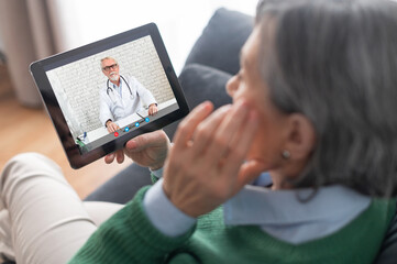 Close-up of a senior mature lady wearing a green sweater holding a tablet and talking with a therapist doctor, getting consultation about her health condition, cyber medicine or self-isolation concept