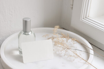 Feminine lifestyle cosmetic still life scene. Glass bottle, flacon with perfume or eau de toilette and dry grass on white marble tray near window. Blank business card mockup. Selective focus.