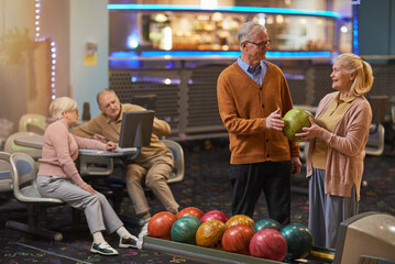 Fototapeta premium Portrait of smiling senior couple playing bowling together with friends in background while enjoying active entertainment at bowling alley, copy space