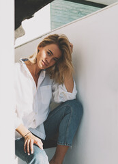 Beautiful smiling blonde woman wearing white shirt and blue jeans looking at the camera adjusting her hair with the hand while sitting at the steps of the beach house staircase on a warm sunny day. - 408163228