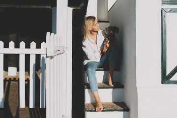 Charming blonde woman in white shirt and blue jeans looking tenderly at her lovely pet dog laying on her knee while sitting barefoot at the stair steps of a beach house on a warm sunny day.