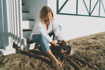 Cheerful blonde woman sitting at the bottom step of the beach house staircase and stroking her cute pet dog on a sunny day. Beautiful Caucasian female having fun with her dog at the sea shore.