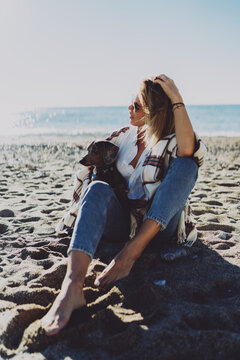 Beautiful model look blonde female smiling while sitting with her lovely dog and relaxing on a beach side. Cheerful woman sitting barefoot on sand with her puppy enjoying sunny day at the seaside