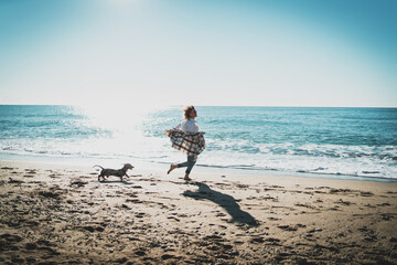 Happy blonde woman running alongside the sea shore with her pet dog on a sunny day. Cheerful Caucasian female in blue jeans with plaid on her shoulders playing with her doggy at the beachside.