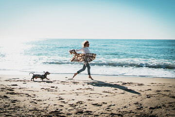 Smiling blonde woman running alongside the sea side with her pet dog on a sunny day. Cheerful Caucasian female in blue jeans with plaid on her shoulders playing with her doggy at the sandy beachside. - 408163675
