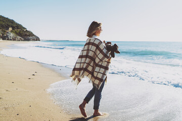 View from the backside photo of a blonde barefoot woman in sunglasses and casual outfit with a plaid on her shoulders walking the seashore with her cute doggy in her arms on a sunny and windy day.