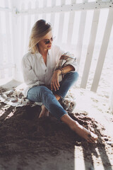 Pensive barefoot blonde woman sitting in the shadow of a wooden pier at the seaside. Charming Caucasian female in sunglasses wearing white shirt and blue jeans sitting at the sandy beach side.