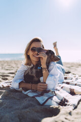 Cheerful female in casual outfit laughing and hugging dog while lying on a sand beach at the day off. Young woman smiling at the camera while relaxing with her pet dog on a warm sunny day outdoors.