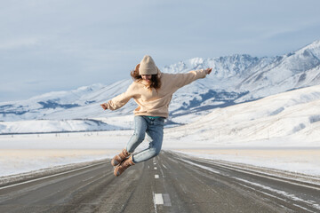 Happy woman jumping on the road against snowy mountains. Young woman in jeans and hoodie enjoys road trip - Powered by Adobe