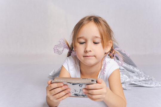A smiling girl with two pigtails in a white dress plays games on the phone, writes text messages, watches a video lying on the floor, isolated on a white background.