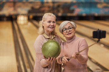 Fototapeta premium Waist up portrait of two senior women taking selfie photos while playing bowling and enjoying active entertainment at bowling alley, copy space