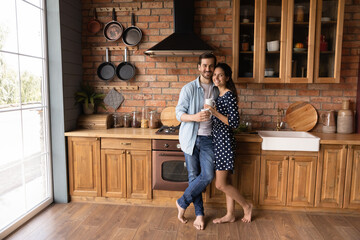 Portrait of happy young Caucasian couple renters or tenants pose in cozy modern renovated design kitchen at home. Smiling millennial man and woman hug embrace enjoy family morning drinking coffee.