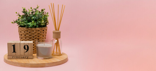 Obraz premium calendar. 19 february composition from a wooden calendar, a flower in a wicker pot, a gray candle in a glass candlestick and an aroma diffuser on a light pink background banner business