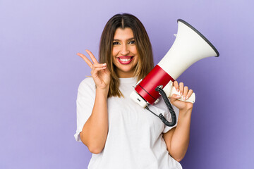 Young indian woman holding a megaphone isolated joyful and carefree showing a peace symbol with fingers.