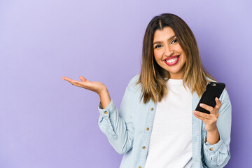 Young indian woman holding a phone isolated showing a copy space on a palm and holding another hand on waist.