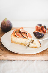 Slice of homemade fig cheesecake with fresh figs on white plate over rustic wooden board, white wall at background, close-up. Healthy dessert, artisan sweet food concept