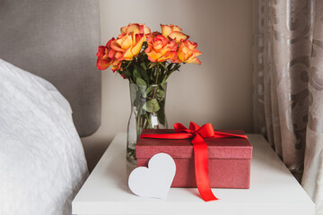 A beautiful gift box with red ribbon, a bouquet of roses flowers in vase and heart shaped blank greeting card on the bedside table against a gray wall. Valentine's day, birthday, women's day surprise.