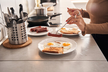 Woman pricks a sausage on a fork over plate with bread and eggs