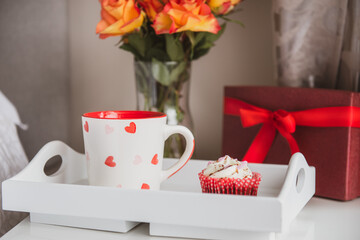 Tray with fresh coffee and cupcake, gift box and a bouquet of roses flowers in vase on the bedside table against a gray wall. Valentine's day, birthday, women's day festive breakfast. Selective focus.
