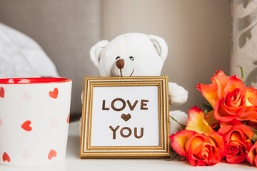 Morning cup of fresh coffee, roses bouquet, white teddy bear with Love you message in frame on the bedside table. Valentine's day, birthday, women's day greeting idea. Love concept. Selective focus