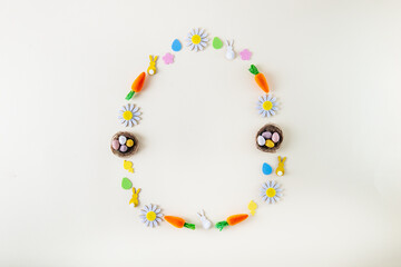Creative Easter. Flat lay with egg's shape,  frame of candy eggs, and another festive decor. Decorative carrots, rabbits, chickens, flowers on the light background. Minimal spring concept. Copy space.