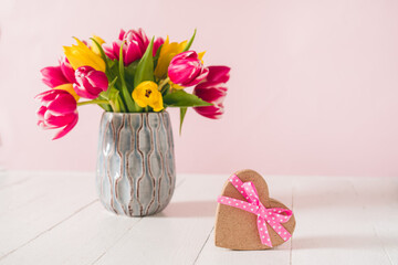Craft heart shape gift box with satin ribbon bow and spring bouquet of pink and yellow tulip flowers in a vase on the white wooden table and pink background. Greeting card mockup. Copy space.