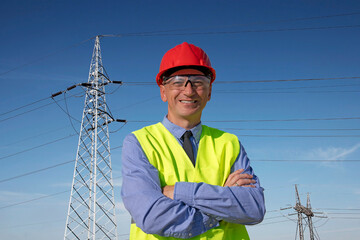 Smiling Engineer in Red Hardhat and Yellow West Standing Under The Electrical Transmission Towers