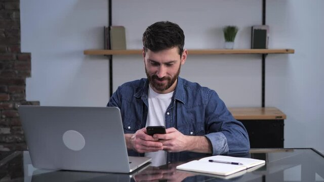 Isolated at home young handsome caucasian man sitting at the work table using laptop computer tapping scrolling browsing typing writing sending message on his smartphone screen. E-commerce remote work