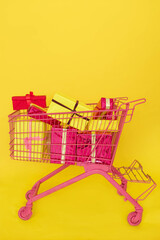 Gift buying. Shopping cart full of boxes. Pink trolley with presents isolated on yellow background. Christmas, Xmas, birthday gift. Concept of celebration, retail and sale. Online Shopping. copy space